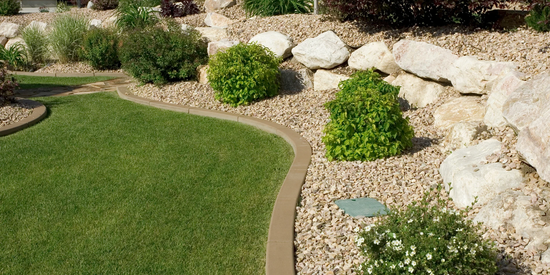 Professionally landscaped yard with curved concrete edging, decorative rock ground cover, large boulders, and healthy green shrubs next to a lush manicured lawn